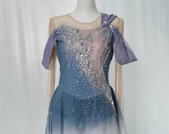 May include: A figure skating costume on a mannequin. The dress features a blue and white ombre skirt, a bodice with silver embellishments, and sheer sleeves. The text "Glamour On Ice" and "Figure Skating Costumes by Chloe" are visible.