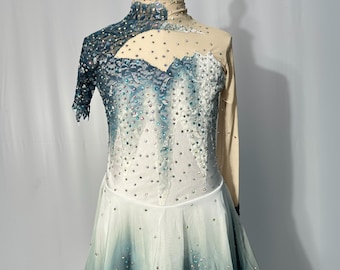 May include: A figure skating costume with a white and blue ombre skirt and a long-sleeved bodice with a lace overlay. The bodice has a sheer design with rhinestones and a blue and white pattern. The costume is on a mannequin and has the text "Glamour On Ice Figure Skating Costume by Chloe" at the bottom.