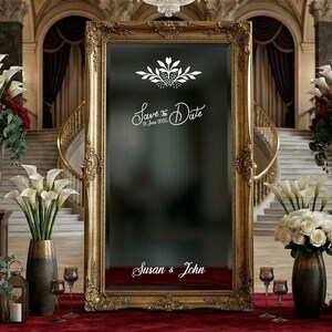 May include: A grand hall decorated for a wedding. A large gold-framed mirror with "Save the Date" text is the focal point. Floral arrangements of red roses, white calla lilies, and greenery are displayed in ornate vases. A red carpet leads to the mirror.