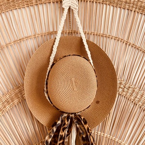 May include: A straw hat with a leopard print ribbon hanging on a macrame hat holder against a woven wall.