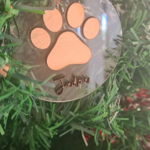 May include: Clear acrylic Christmas ornament with a wooden paw print design and the name "Jadlow" in a cursive font. The ornament hangs from a natural twine string and is set against a backdrop of a green Christmas tree.