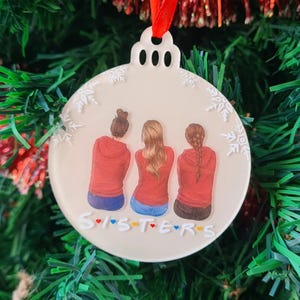 May include: A cream-colored Christmas ornament with a red ribbon. The ornament features an illustration of three figures in red hoodies and blue jeans, with the word "SISTERS" in colorful letters below. White snowflake accents decorate the edge.