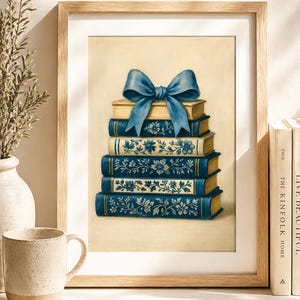Op de afbeelding: Een ingelijste print met een stapel boeken met blauwe en gouden covers, bekroond met een grote blauwe strik. De boeken hebben bloemmotieven. Het kunstwerk bevindt zich in een houten lijst en wordt gepresenteerd op een wit oppervlak.