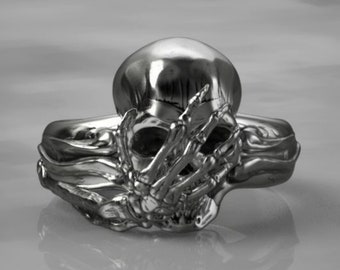 May include: A silver ring with a skull design. The skull is covering its eyes with its skeletal hand.
