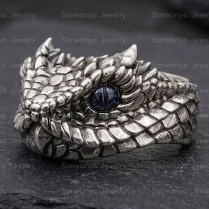 May include: A silver ring shaped like a snake head with detailed scales and a single dark blue eye. The metal has an oxidised finish that highlights the texture of the serpent skin. The design features a pointed snout and small horn-like protrusions.