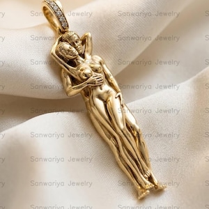 Può includere: Un ciondolo color oro con un design scolpito di due figure che si abbracciano. Il gambo è adornato con piccole pietre preziose trasparenti. Il ciondolo è esposto su un tessuto color crema strutturato.