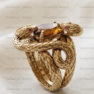 May include: A gold ring with a design of two snakes entwined around a central amber-coloured gemstone. The snakes have ruby eyes. The ring's intricate design and textured finish are set against a neutral background.
