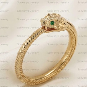 Könnte beinhalten: Ein goldfarbener Armreif in Schlangenform mit einem strukturierten, schuppenartigen Muster entlang des Bandes. Der Schlangenkopf ist mit einem kleinen, leuchtend grünen Edelstein als Auge verziert und das Maul ist leicht geöffnet.