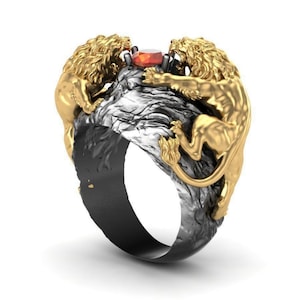 Può includere: Un anello d'oro con due leoni che si fronteggiano, tenendo una pietra preziosa rossa tra le loro zampe. L'anello è montato su una fascia nera e argentata.