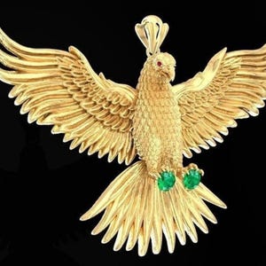 Könnte beinhalten: Ein goldfarbener Anhänger in Form eines Vogels mit ausgebreiteten Flügeln. Der Vogel hat smaragdgrüne Edelsteine an seinen Füßen.