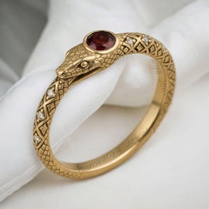 Op de afbeelding: Een goudkleurige ring in de vorm van een slang. De kop van de slang heeft een ronde, rode edelsteen. Het lichaam van de ring is getextureerd met ruitvormige patronen en kleine, heldere edelstenen. De ring wordt op een wit oppervlak getoond.
