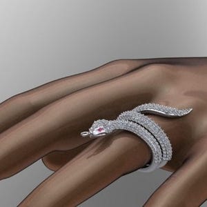 May include: A silver snake ring with a detailed design. The ring features a snake shape with a head and tail, and is encrusted with small, sparkling stones. The snake's eye is a small, red stone. The ring is worn on a finger.