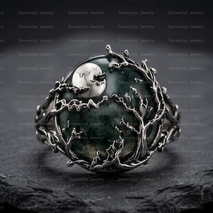 May include: A silver ring featuring a round, dark green stone centerpiece. The metal band is crafted into a detailed, raised design of bare, gnarled tree branches that frame the stone, with a small, polished silver moon accent at the top edge.