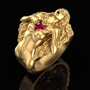 Peut inclure: Une bague en or représentant une silhouette sculptée aux longs cheveux ondulés, les mains dirigées vers la poitrine où est sertie une pierre précieuse rouge vif à facettes. La figure présente une bouche ouverte et des traits détaillés.