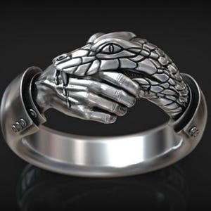 May include: A silver ring featuring a detailed design of a snake wrapped around a human hand. The snake's scales and the hand's fingers are intricately sculpted.