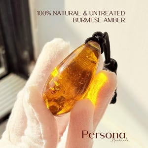 Könnte beinhalten: Ein tropfenförmiger Anhänger aus durchscheinendem goldbraunem burmesischem Bernstein, der an einer schwarzen Kordel hängt. Der Text 100% NATURAL & UNTREATED BURMESE AMBER und Persona Handmade ist sichtbar. Der Bernstein zeigt Einschlüsse und Lichtbrechung.