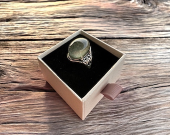 May include: A silver ring featuring an oval-shaped, shimmering dark green gemstone set in a decorative metal band. The ring is displayed inside a square, light-colored cardboard gift box with a black velvet insert and a small fabric pull tab.