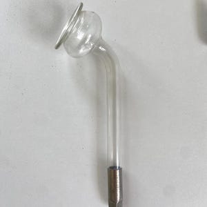 Puede incluir: Un electrodo de alta frecuencia de vidrio transparente con cuello curvo y base metálica. El electrodo tiene una parte superior redondeada con un borde circular. La base es de color plata mate.