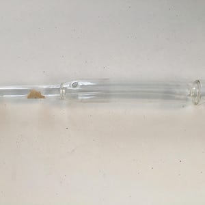 Puede incluir: Un tubo de vidrio transparente con un extremo redondeado y un pequeño agujero cerca del centro. El tubo mide aproximadamente 30 cm de largo.