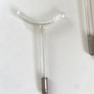 Puede incluir: Varita de vidrio transparente con una parte superior curva y un tallo recto. El tallo tiene una base de metal. La varita es probablemente parte de un dispositivo o herramienta más grande. La imagen está tomada sobre una superficie blanca.