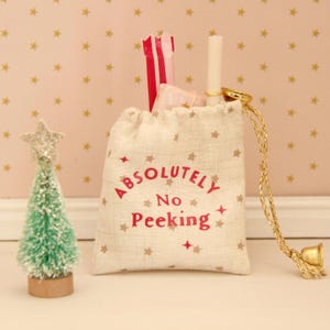 May include: A small beige drawstring pouch featuring a star pattern and the red text "ABSOLUTELY NO PEEKING" printed on the front. The bag is cinched with gold cords and contains a white tube and a red and white striped item, next to a small green tree.