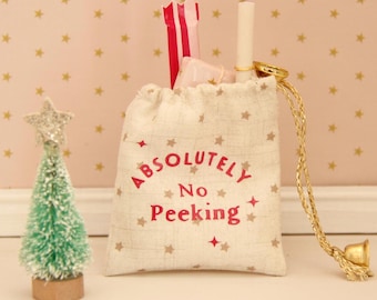 May include: A small beige drawstring pouch featuring a star pattern and the red text "ABSOLUTELY NO PEEKING" printed on the front. The bag is cinched with gold cords and contains a white tube and a red and white striped item, next to a small green tree.