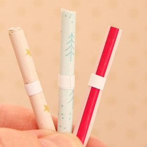 May include: Three cylindrical paper scrolls secured with white paper bands. The left scroll is cream with gold stars, the centre scroll is light blue with dark blue pine tree patterns, and the right scroll features a bold vertical red stripe design.