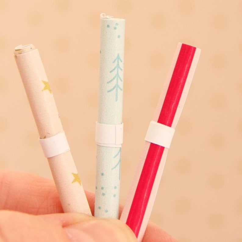 May include: Three cylindrical paper scrolls secured with white paper bands. The left scroll is cream with gold stars, the centre scroll is light blue with dark blue pine tree patterns, and the right scroll features a bold vertical red stripe design.