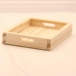 May include: A small, rectangular, light-coloured wooden tray with a shallow base and low walls. The tray features two cut-out handles on opposite sides, one on the front and one on the back, and displays a natural wood grain texture throughout.