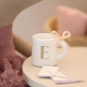 May include: A miniature white mug with a gold letter "E" on the front, adorned with a light pink bow. Two small white envelopes and a pink pencil are in front of the mug. The mug is on a light pink surface.