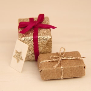 May include: A square gift box wrapped in gold glitter paper with a dark pink ribbon tied in a bow, a rectangular gift box wrapped in brown kraft paper with a thin gold twine bow, and a small white card featuring a gold glitter star.