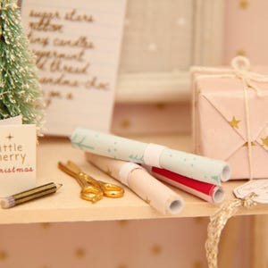 May include: A miniature scene featuring a small bottle-brush tree, a tiny card with the text 'little merry christmas', a gold-coloured pencil, gold-toned scissors, three rolled sheets of patterned wrapping paper, and a gift box with gold stars.