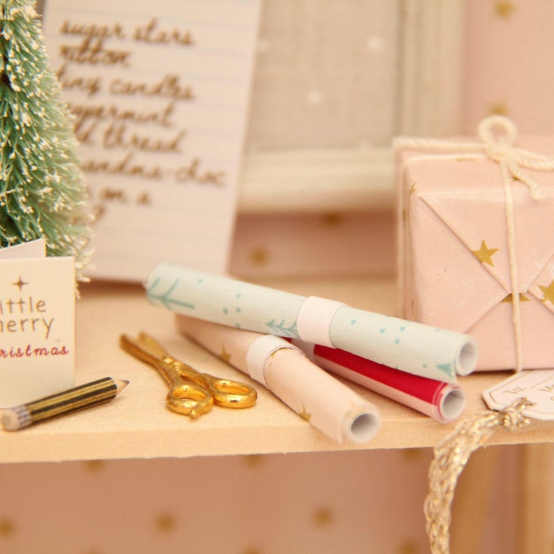 May include: A miniature scene featuring a small bottle-brush tree, a tiny card with the text 'little merry christmas', a gold-coloured pencil, gold-toned scissors, three rolled sheets of patterned wrapping paper, and a gift box with gold stars.