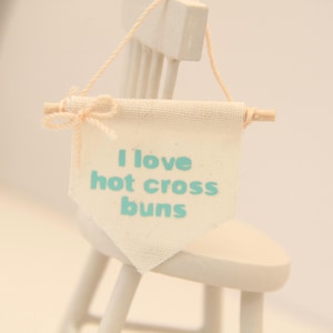 May include: A miniature banner in off-white fabric, reading "I love hot cross buns" in light blue. It's suspended from a wooden dowel with a small bow. The banner hangs on a small white chair, a charming decorative item.