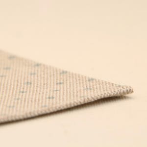 May include: A piece of beige fabric with a woven texture featuring a repeating pattern of small, light blue dots. The fabric is folded at an angle, showing the thickness of the material and the consistent weave of the textile fibres throughout.