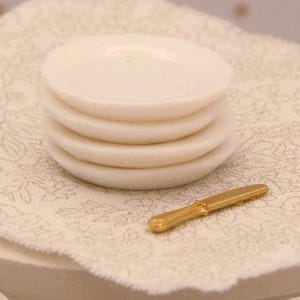 May include: A stack of four small, round, off-white ceramic plates with smooth, slightly raised edges. Beside the stack of plates lies a single, small, metallic gold-coloured knife with a textured handle and a serrated blade edge.