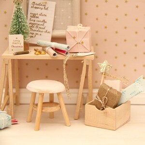 May include: A miniature wooden desk and stool set featuring a small Christmas tree, a list titled Christmas shopping list, wrapped gifts, and a wooden crate containing a brown parcel and a light blue patterned paper roll with gold accents.