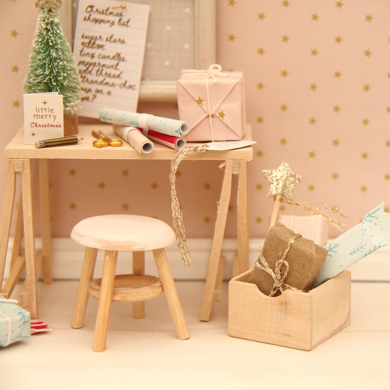 May include: A miniature wooden desk and stool set featuring a small Christmas tree, a list titled Christmas shopping list, wrapped gifts, and a wooden crate containing a brown parcel and a light blue patterned paper roll with gold accents.