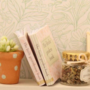 May include: Miniature garden-themed decor featuring two small books titled "The little greenhouse book" and "The little garden book." A small terracotta pot with a succulent plant and a glass jar filled with herbs complete the scene.