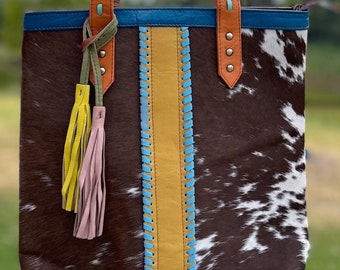 May include: A brown and white cowhide tote bag with a blue, yellow, and orange trim. The bag has two long straps and a tassel.