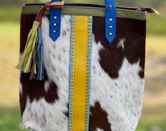 May include: A brown and white cowhide tote bag with yellow, blue, and green trim. The bag has a zipper closure and two top handles with blue and red stitching.