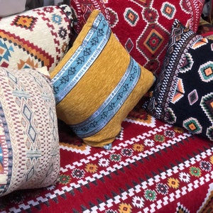Peut inclure: Plusieurs coussins décoratifs aux motifs géométriques variés. Les couleurs incluent le rouge, le jaune, le noir et le crème, avec des motifs et des textures complexes. Les coussins sont disposés sur une surface rouge à motifs.