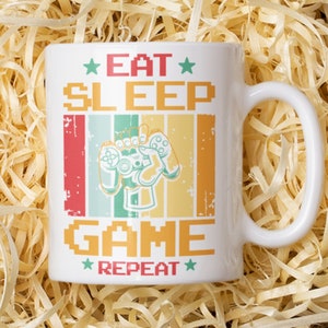 May include: White ceramic mug with a retro-style design featuring a pixelated graphic of a hand holding a video game controller. The text on the mug reads "Eat Sleep Game Repeat" with stars on either side of the text.