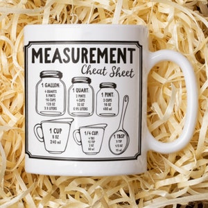 May include: White ceramic mug with a black and white "MEASUREMENT Cheat Sheet" design. The mug features illustrations and measurements for gallons, quarts, pints, cups, and tablespoons in ounces, cups, and liters.