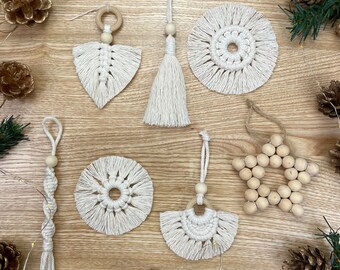 May include: A set of six handmade macrame ornaments in white with wooden beads and rings. The ornaments are displayed on a wooden surface with pine cones and string lights.