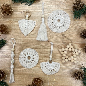 Op de afbeelding: Een set van zes handgemaakte macramé-versieringen in wit met houten kralen en ringen. De versieringen worden getoond op een houten oppervlak met dennenappels en lichtslingers.