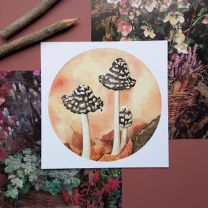 May include: Watercolour painting of three mushrooms with black and white spots on their caps. The mushrooms are painted on a light orange background with brown leaves.