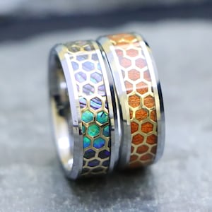 May include: Two silver and gold wedding bands with a honeycomb pattern. One band has a blue and green abalone shell inlay, and the other has a brown wood inlay.