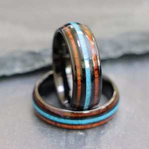May include: Two black tungsten carbide rings with inlaid wood and turquoise. The rings are stacked on top of each other.