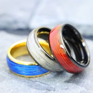 May include: Three men's wedding bands with different colored inlays. The first band is gold with a blue inlay, the second is silver with a white inlay, and the third is black with a red inlay.